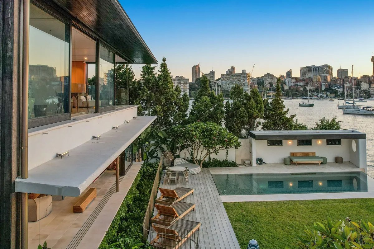 A modern house with large glass windows overlooking a waterway with sailboats, featuring a pool, outdoor seating, and a deck area surrounded by trees and green lawn.