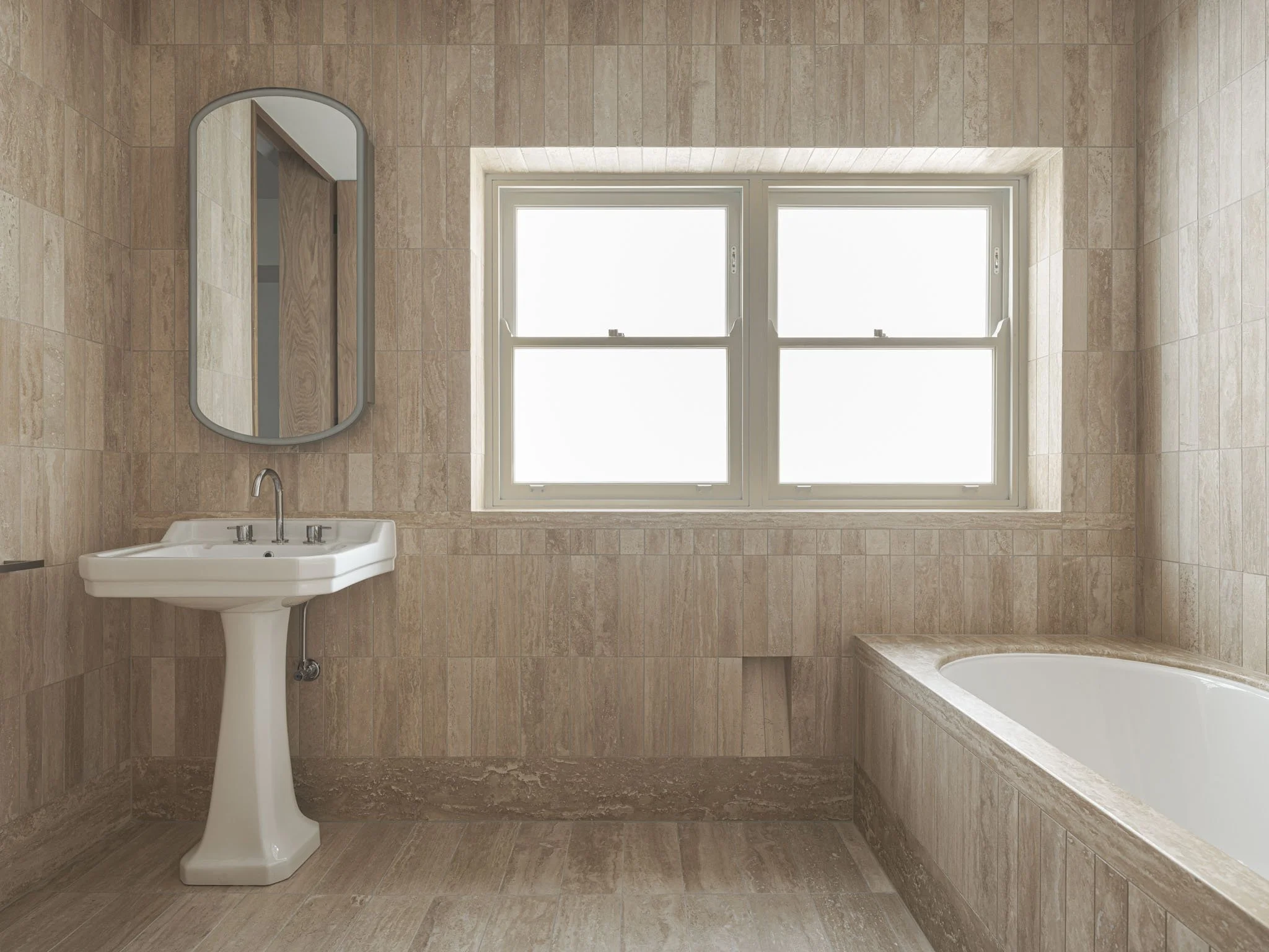 A bathroom with beige tiled walls and floor, featuring a white pedestal sink with a mirror above it, a window with four squares in the background, and a corner bathtub.