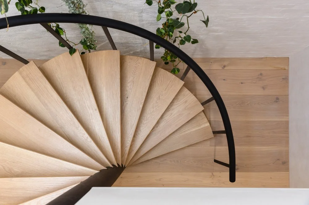 Top-down view of a wooden spiral staircase with black railing, leading down to a wooden floor surrounded by green plants.
