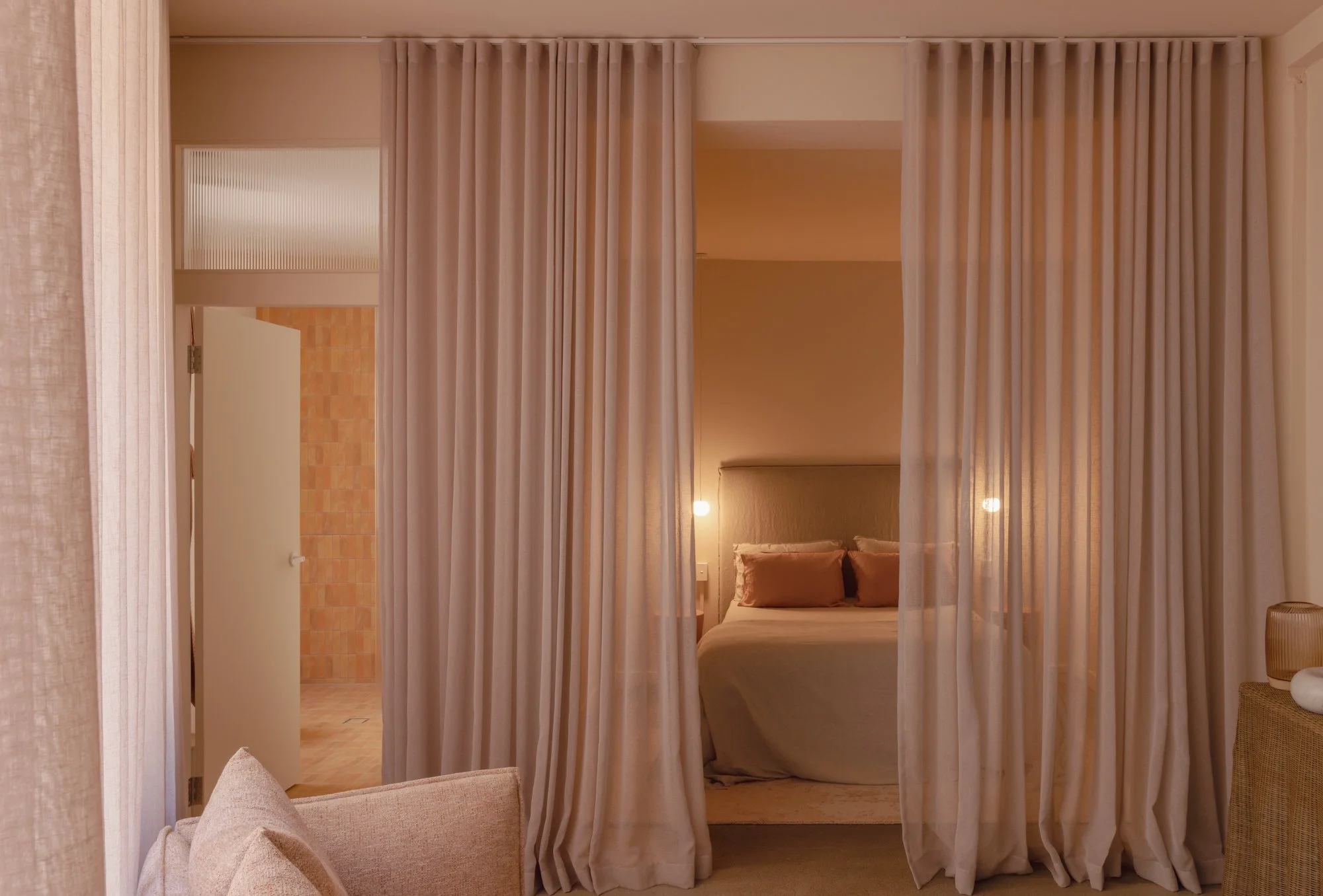 View of a bedroom with beige curtains, a bed with beige linens, and two bedside lamps, seen through a partially closed curtain. An open door reveals a bathroom with peach-colored tiles.