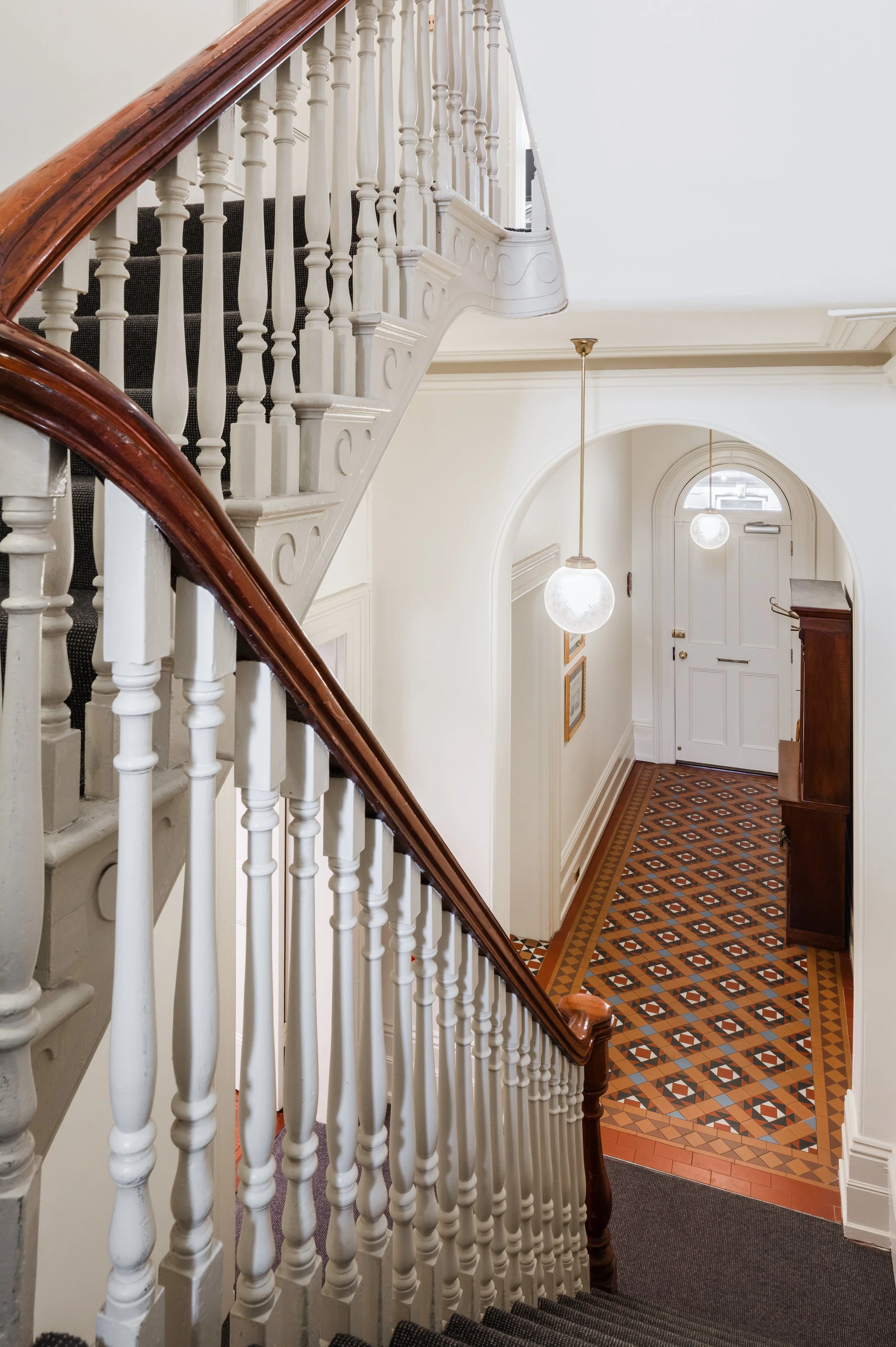 Inside view of a house with a staircase and a tiled hallway. The staircase has a wooden handrail and white balusters. The front door is visible at the end of the hallway, with two hanging globe lights illuminating the space.