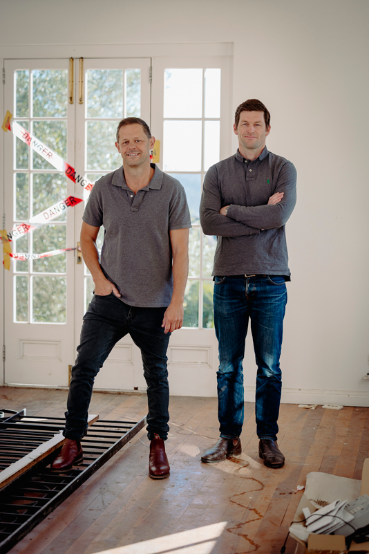 Two men standing inside a room with sunlight coming through large windows. One is standing on a bed frame, both are smiling. There is caution tape hanging in the background.