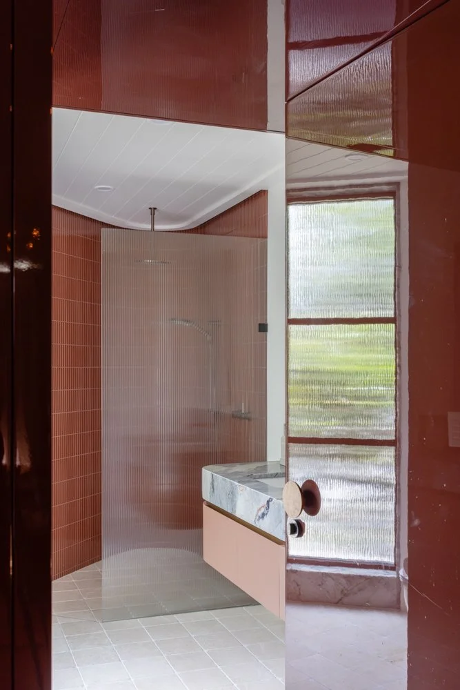 Modern bathroom with brown tiled walls, a curved white ceiling, and a walking shower with clear glass. The bathroom features a marble countertop with a sink, a round brown doorknob, and large windows with silver blinds allowing natural light inside.