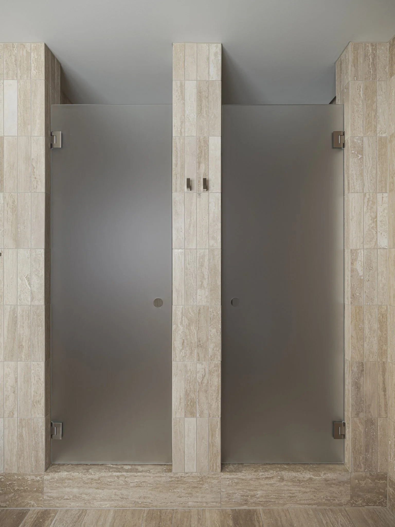 Two closed restroom doors made of gray metal, with beige tiled walls around them.