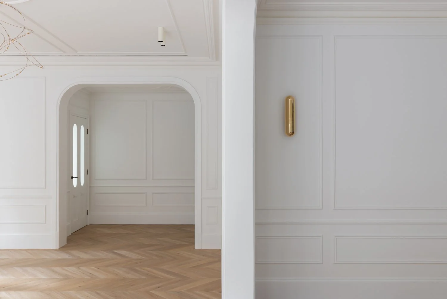 Interior view of a minimalist, white wall with decorative molding, a gold wall-mounted sconce, and a wooden floor in a modern home.