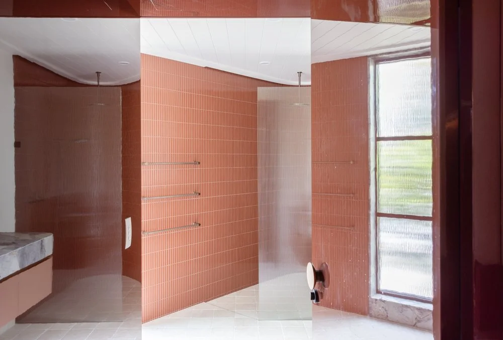 Empty public shower area with brown tiled walls, a large window, and a marble bench.