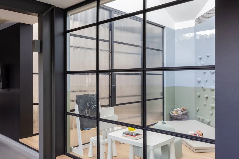 Indoor playroom with large glass window, climbing wall with holds, bean bag, and small table with chairs.