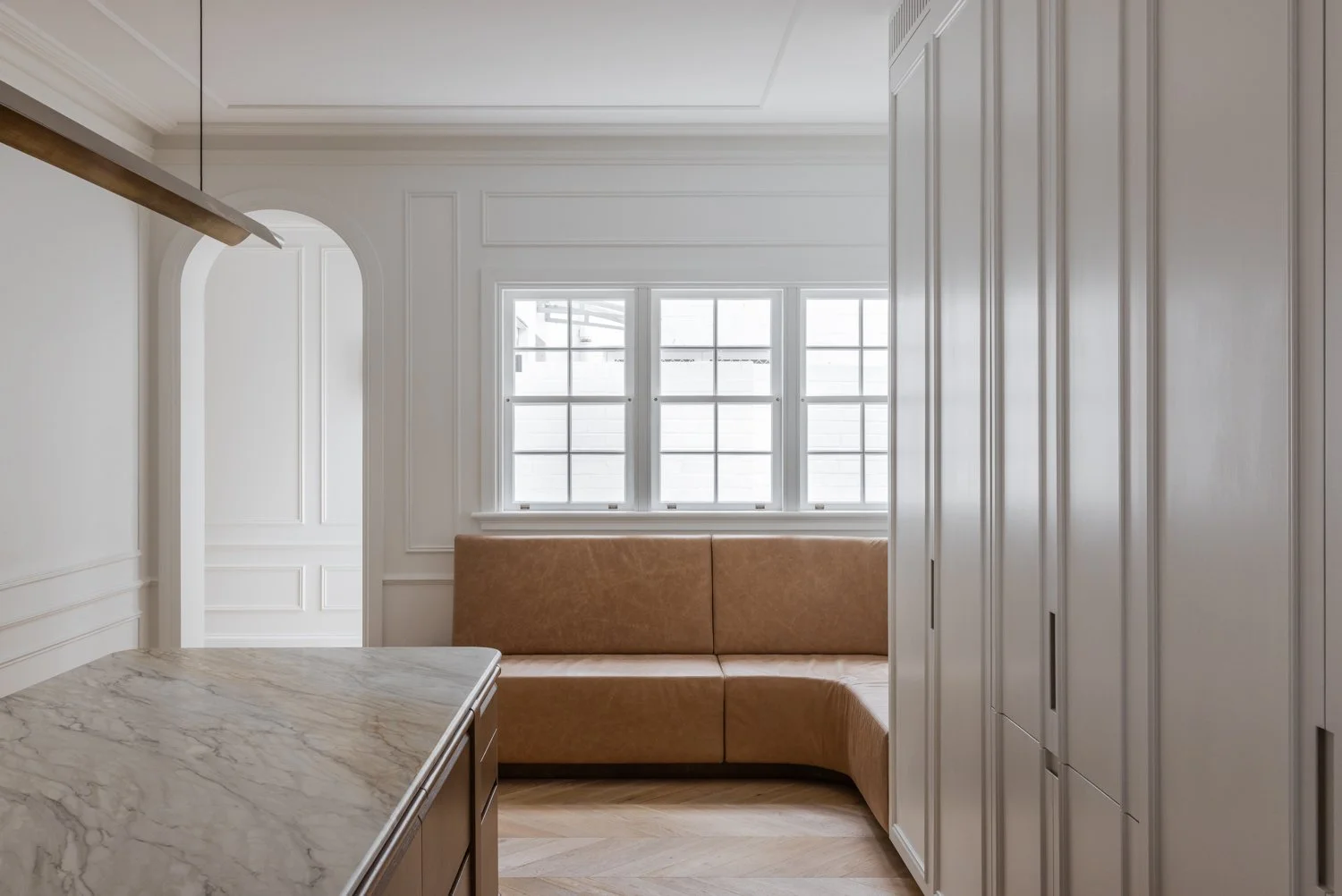Interior view of a modern kitchen or living space with white walls, a marble countertop, and a tan leather built-in bench along the windowed wall.