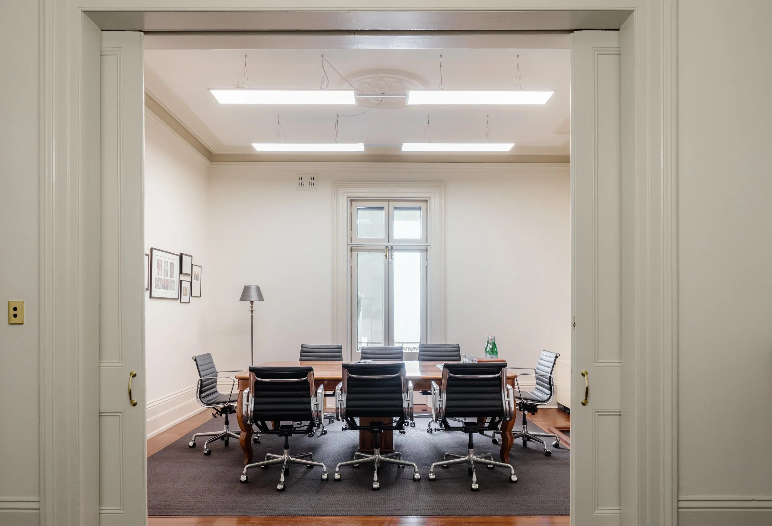 Empty conference room with a wooden table surrounded by eight black leather chairs, a window at the far wall, an adjustable floor lamp, framed pictures on the side wall, and bottled water on the table, with bright ceiling lights overhead.