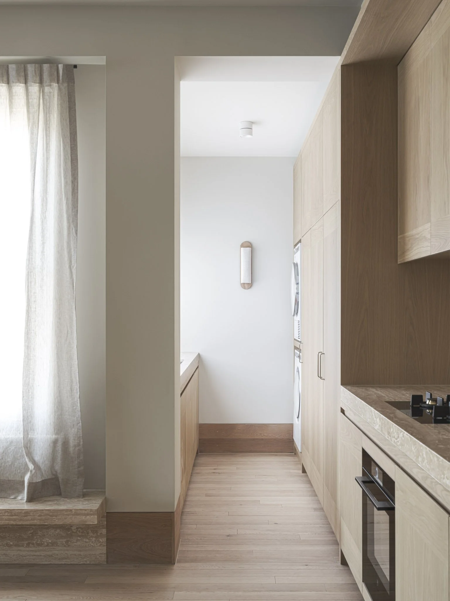 Minimalist kitchen with light wood cabinetry, a marble countertop, and a small kitchen appliance built into the counter. Light wood flooring and a wall-mounted light fixture on a white wall are visible.
