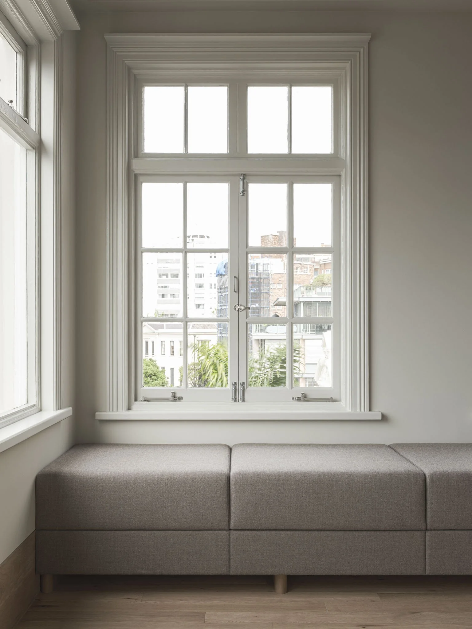 A beige window seat beneath a large white-framed window with multiple panes, looking out onto a cityscape with buildings and trees.