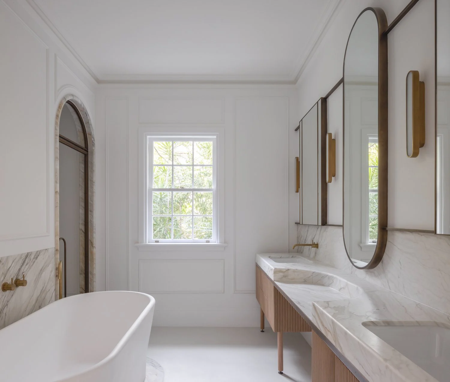 Modern bathroom with a white freestanding bathtub, marble countertops, large framed window, and gold accents.