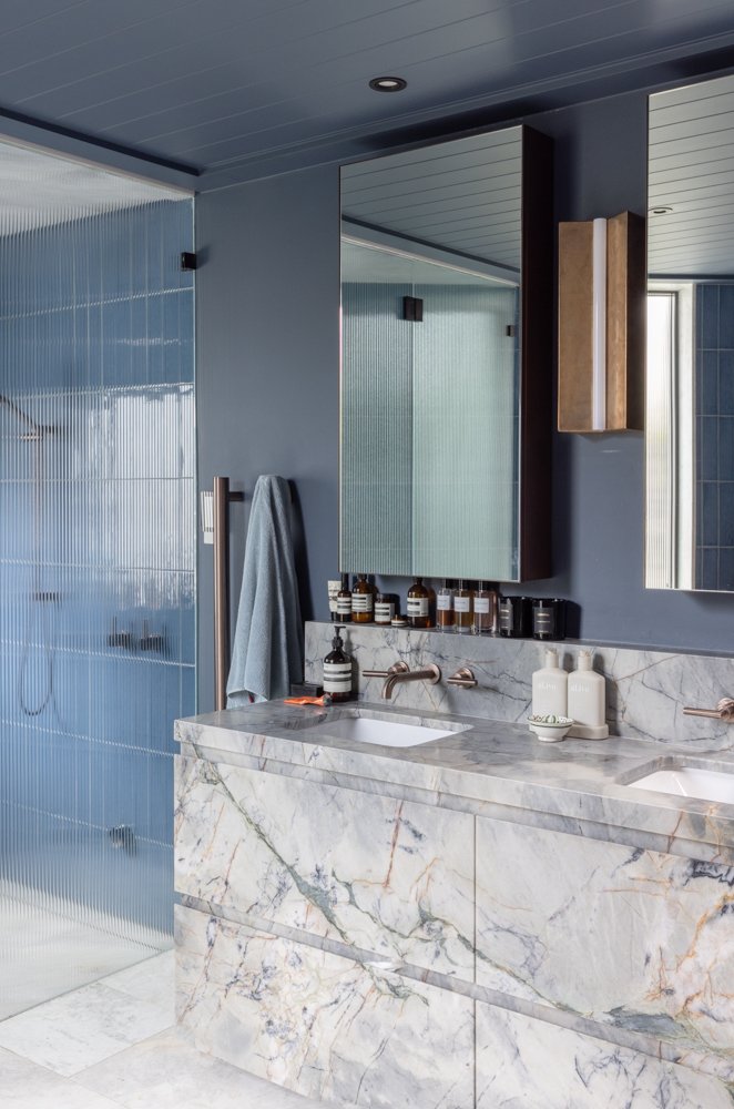 Modern bathroom with marble vanity, a double sink, and dark blue walls. Mirror cabinets and toiletries are visible. Shower with frosted glass door is on the side.
