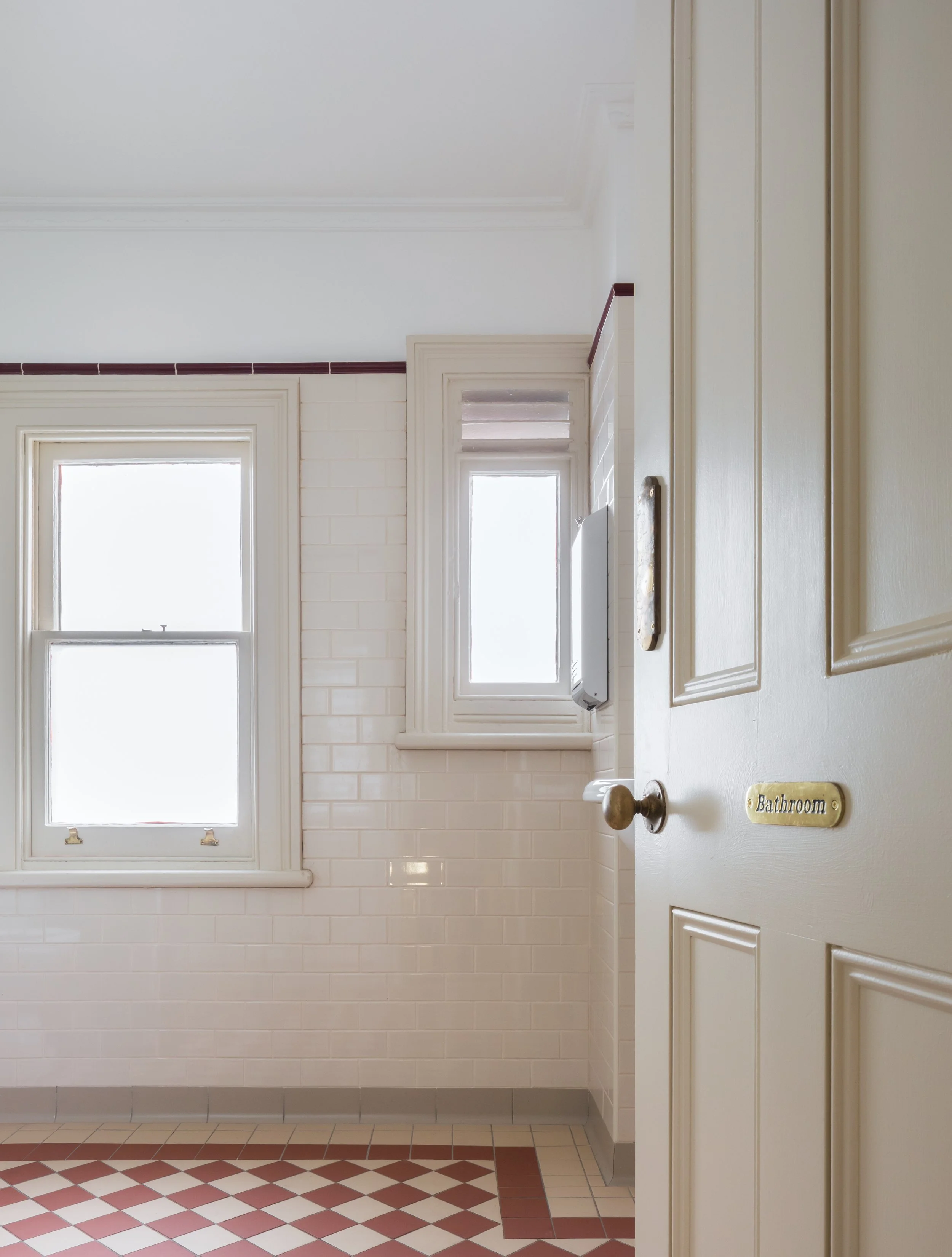 Photo of a bathroom with two windows, white tiled walls, and a door labeled 'Bathroom' slightly open.