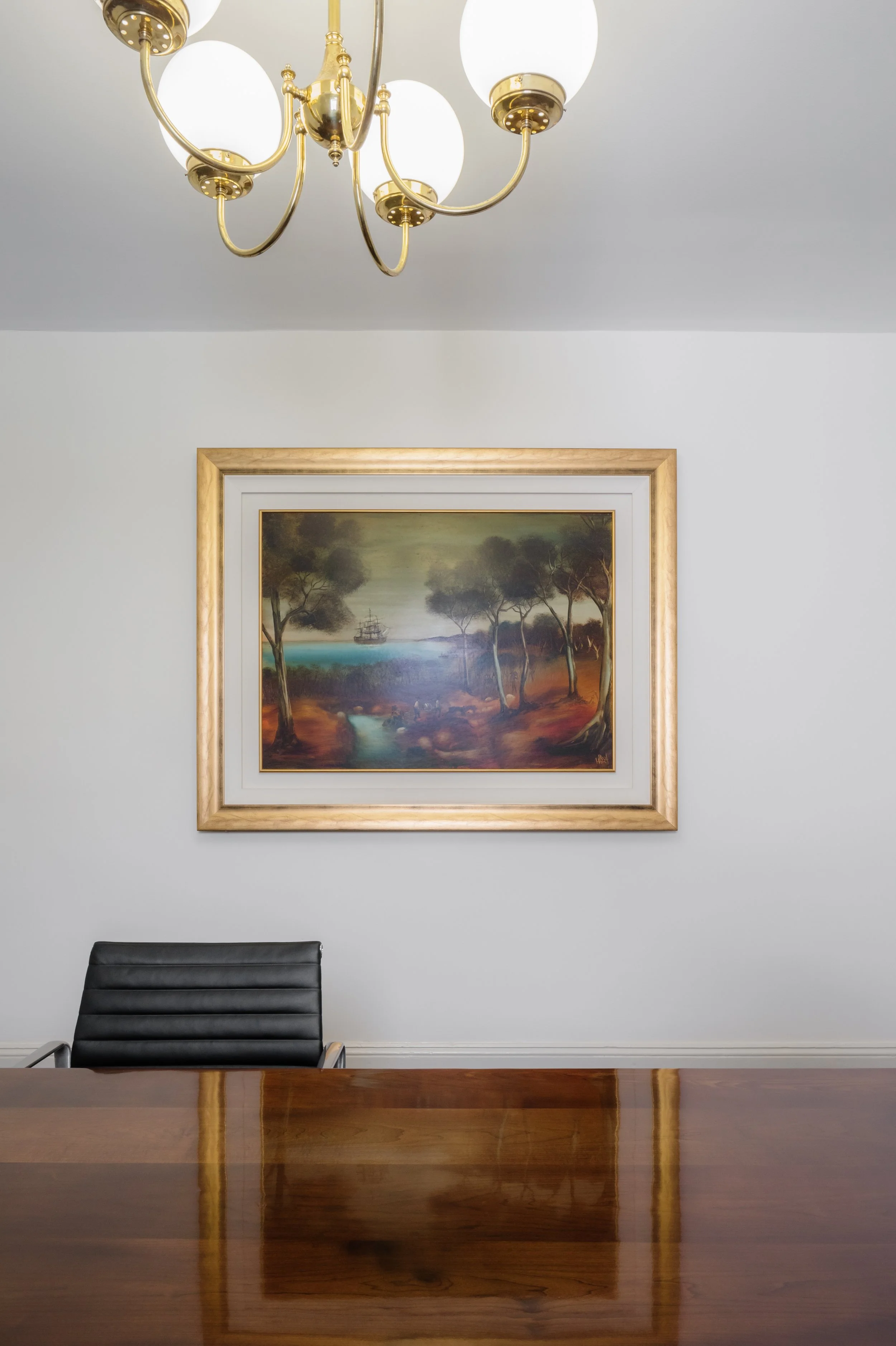 A meeting room with a wooden table, a black leather chair, a framed landscape painting of trees and a boat, and a gold chandelier with white globe lights hanging from the ceiling.