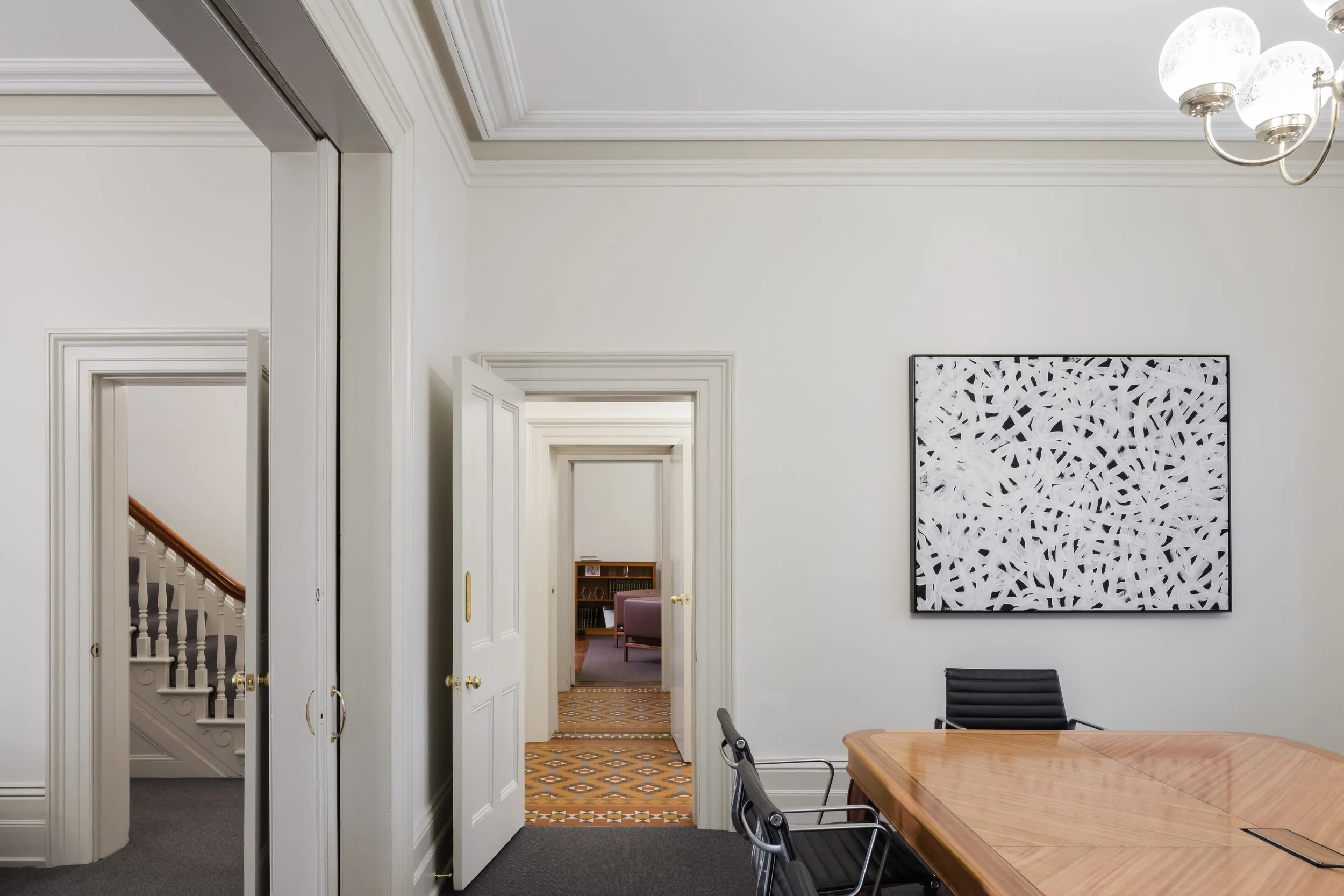 Empty conference room with a wooden table, black chairs, white walls, artwork, and open doors leading to another room with patterned floor and furniture.