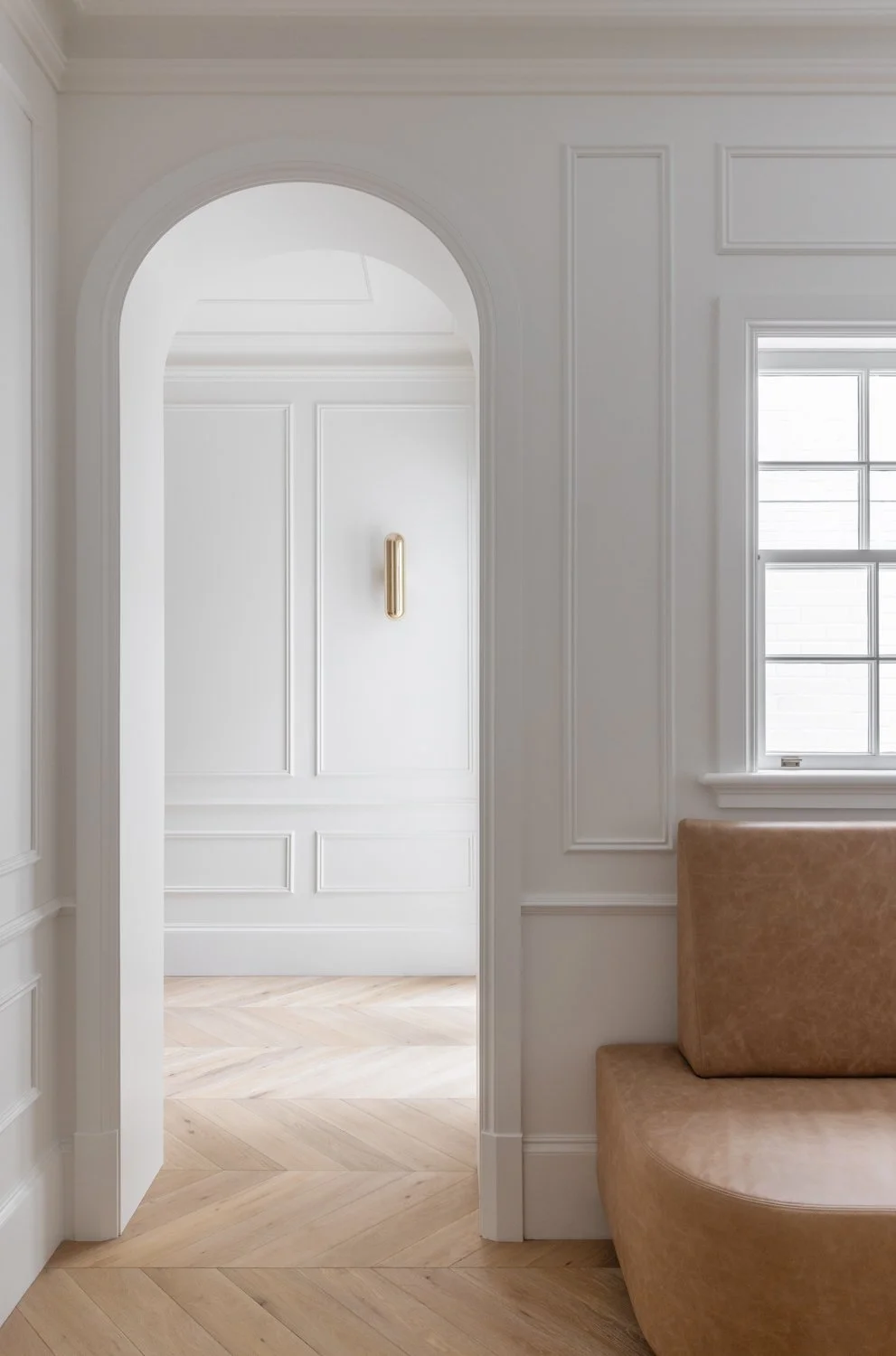 Interior view of a white room with wooden flooring, an arched doorway with a gold handle, and a large window with grid panes. Part of a tan sofa is visible on the right side.