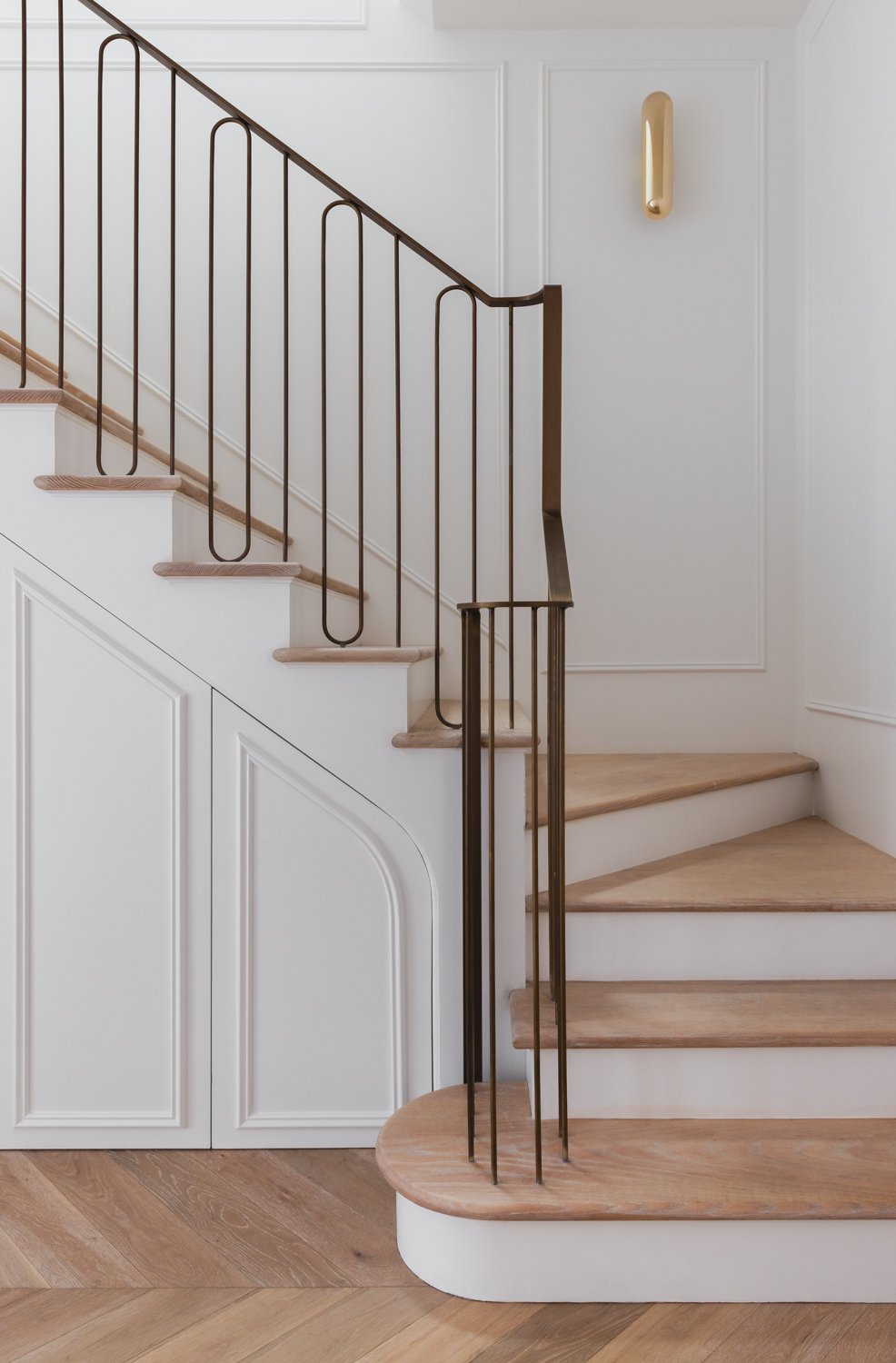 Interior staircase with wooden steps, white panel walls, and a modern metal railing, featuring a gold wall sconce.
