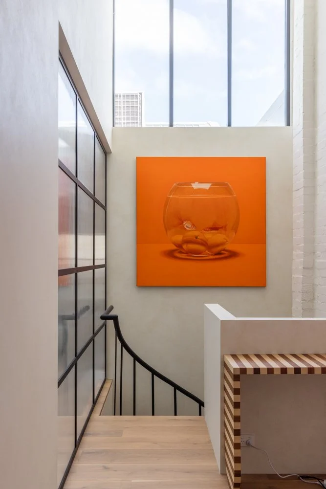 Interior view of a modern stairwell with a large orange painting of a fishbowl hanging on the wall, high windows letting in natural light, and a carved wooden table with a striped pattern.