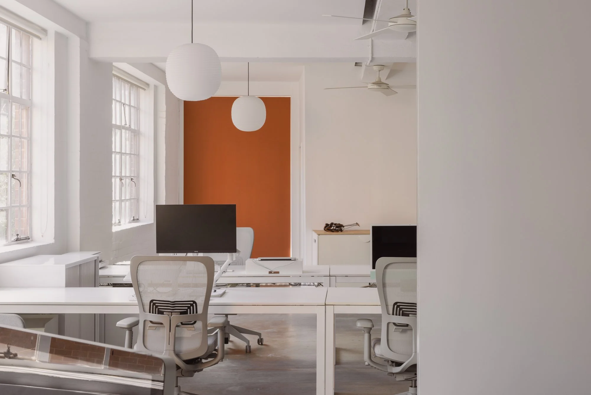 Empty modern office space with white desks, white chairs, a computer monitor, large windows, and a white wall with an orange accent in the background.