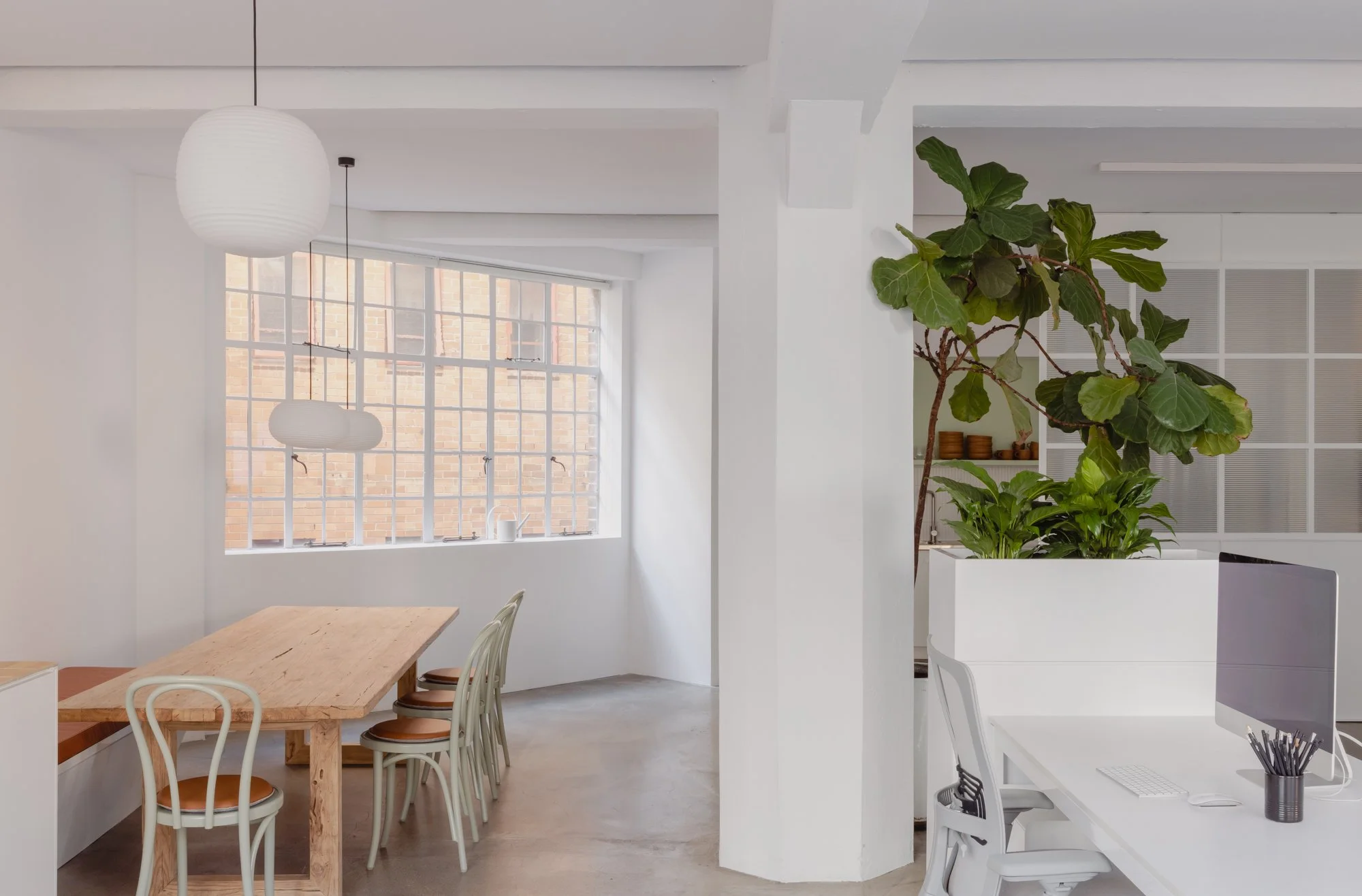 Modern minimalist office interior with a large window, wooden dining table, white chairs, hanging paper lantern lights, potted plants, and a white desk with a computer.