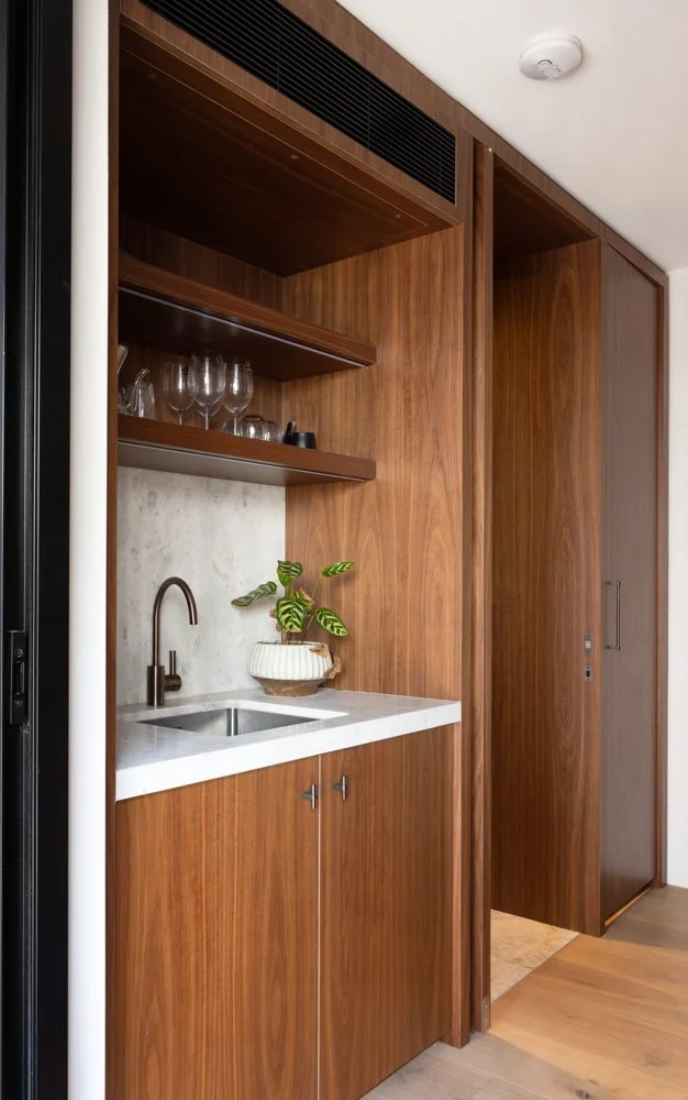 Small kitchenette with a white countertop, a small sink with a bronze faucet, wooden cabinets, two open shelves with glassware, and a potted plant, adjacent to a doorway.