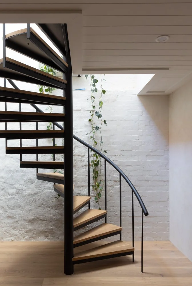 Indoor spiral staircase with black metal railing, wooden steps, white brick wall with a hanging vine plant, and light-colored wooden floor.