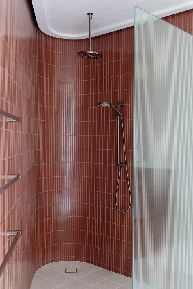 A modern shower with red tile walls, a ceiling-mounted rain shower head, a handheld shower head attached to the wall, a frosted glass partition, and metal grab bars.