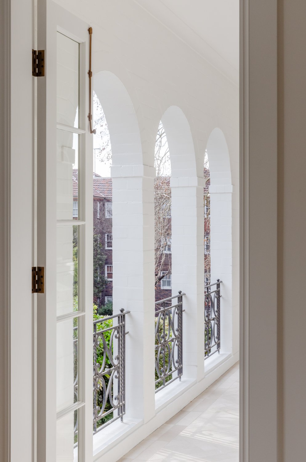 View of a white balcony with arched openings and wrought iron railings, seen from inside a room with open white French doors.