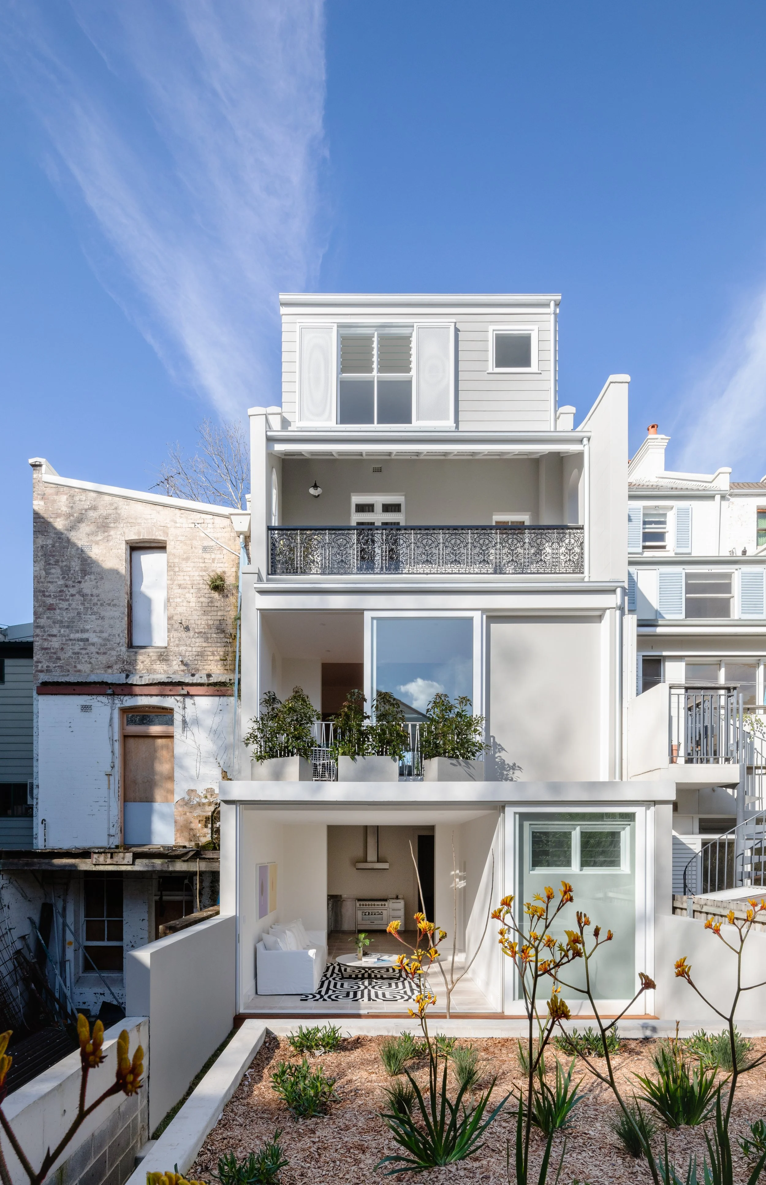 Multi-story modern townhouse with white exterior, balconies, and glass doors, surrounded by a small garden with plants and flowers, under a blue sky.