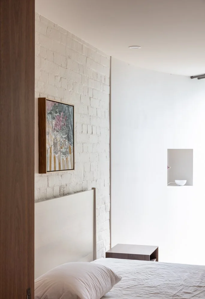 A bedroom with a white brick wall, partial view of a bed with white sheets and pillow, a small dark bedside table, a framed painting on the wall, and a small wall shelf with a white bowl.
