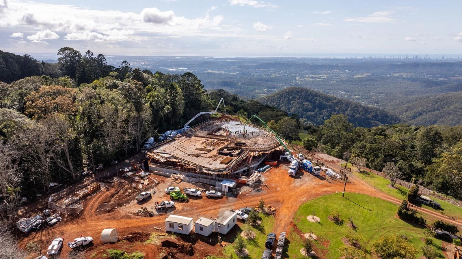 Aerial view of a construction site on a hillside with a partially built structure, construction vehicles, and workers, surrounded by trees with a city skyline in the distance.