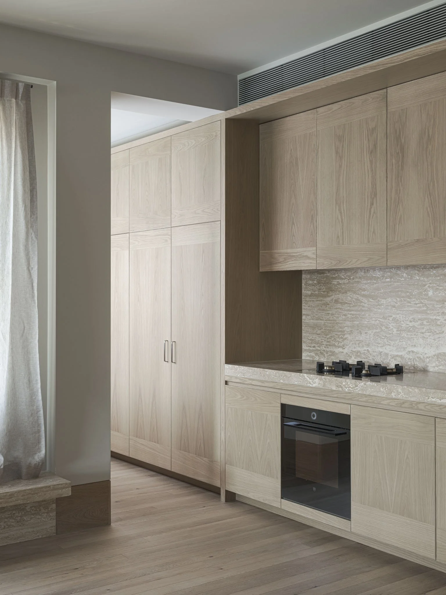 Modern kitchen with light wood cabinets, a built-in oven, gas cooktop, and marble backsplash. There is a curtain on the left side and wooden flooring.