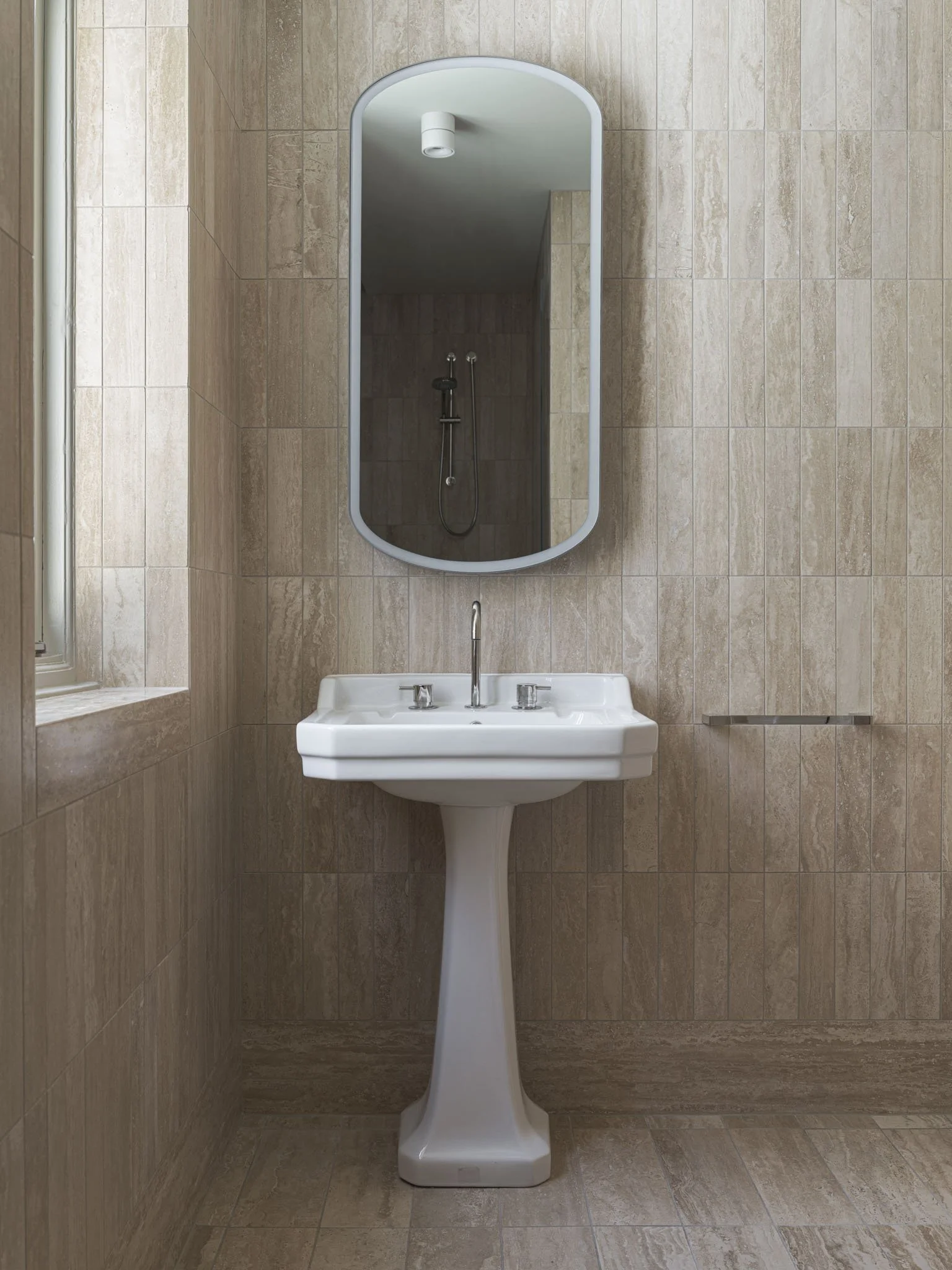 A beige tiled bathroom with a white pedestal sink, a tall oval mirror above, a small window to the left, and a shower with a handheld showerhead reflected in the mirror.