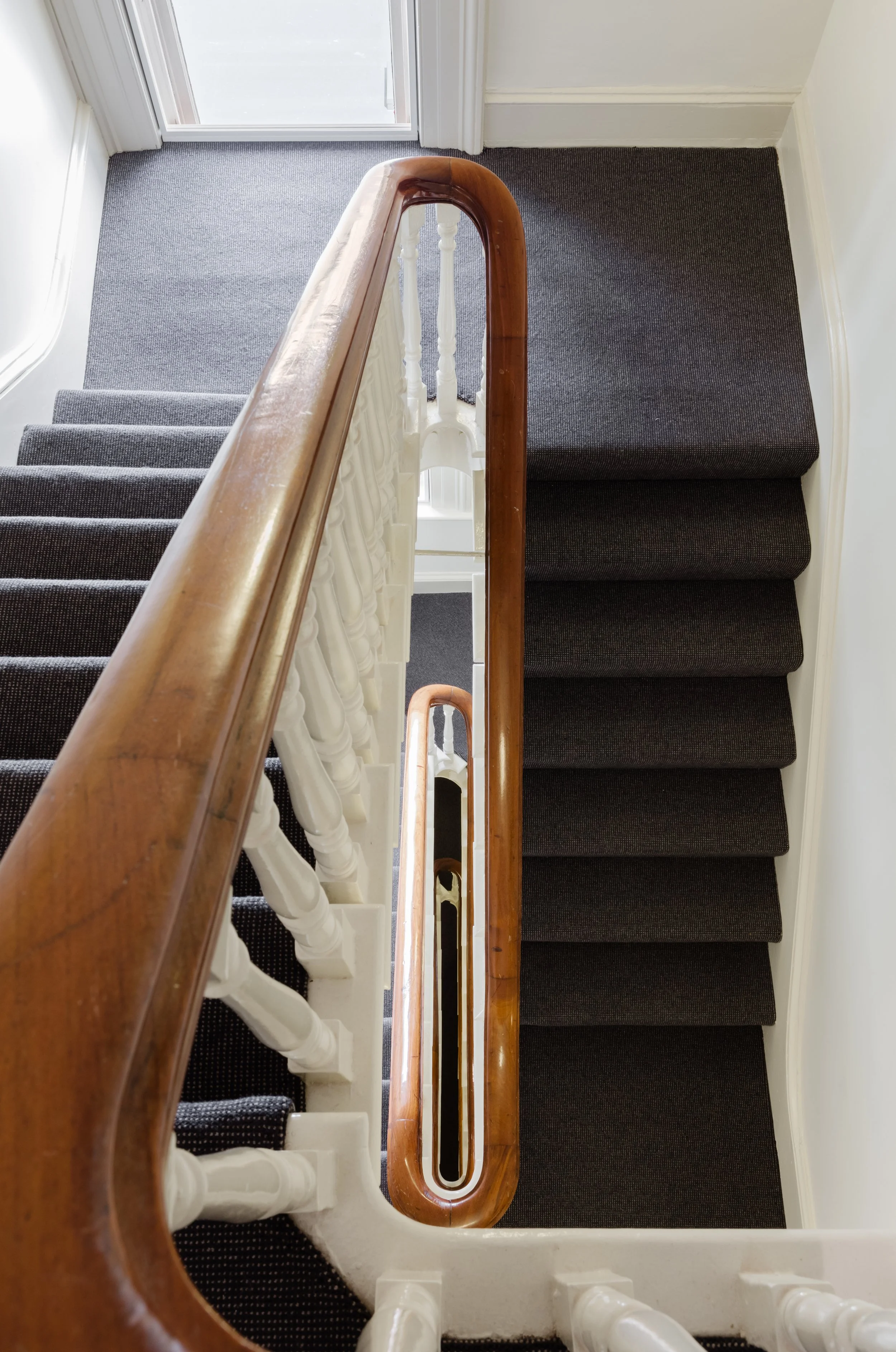 Top view of a staircase with dark carpeted steps, white balusters, and a polished wooden handrail.