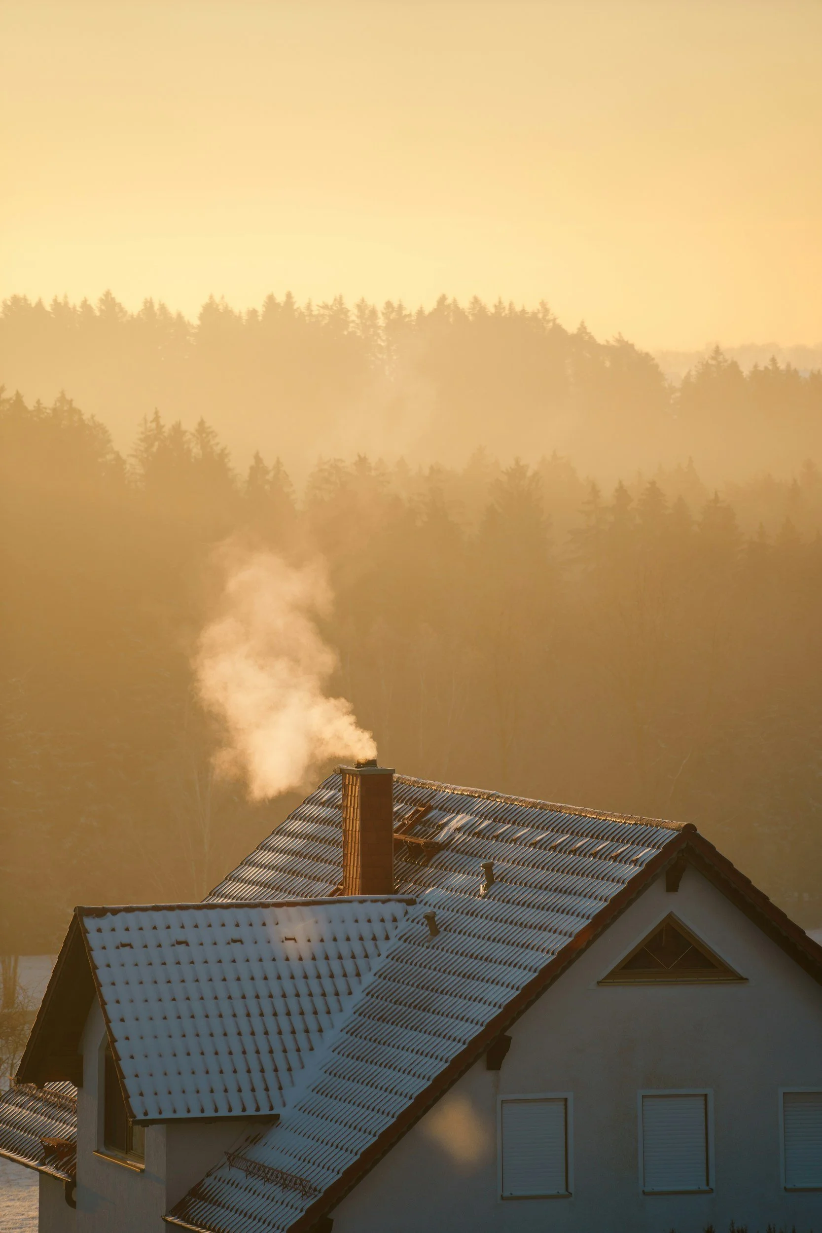 Ein Haus mit schneebedecktem Dach, Rauch steigt aus einem Schornstein, im Hintergrund ein Wald bei Sonnenuntergang.