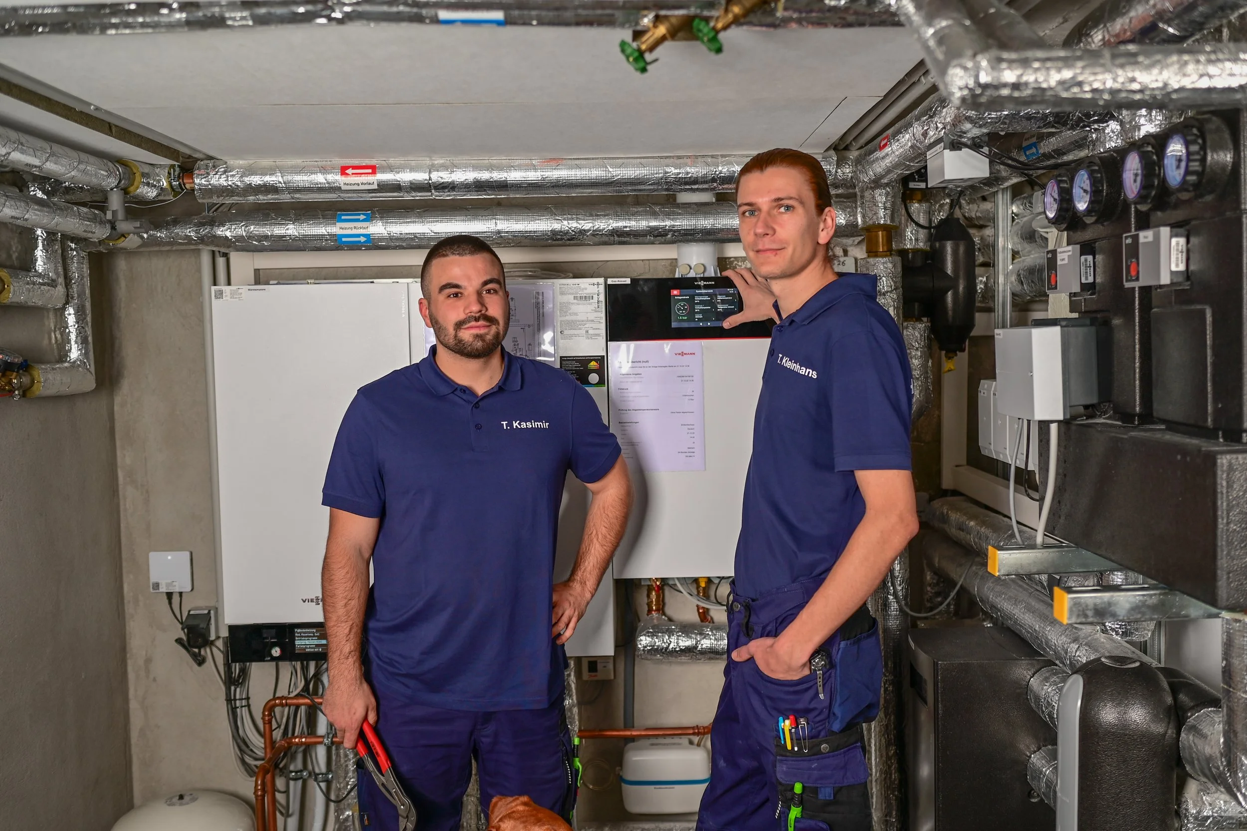 Zwei Techniker in blauen Uniformen arbeiten in einem Heizungsraum mit Geräten, Rohren und einem Display. Einer hält Werkzeuge in der Hand, der andere zeigt auf ein Display.