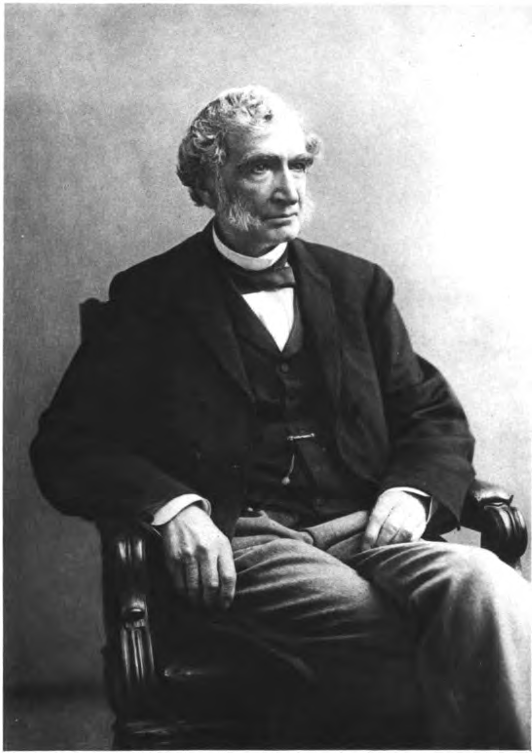 Black and white portrait of an elderly man with wavy gray hair, seated in a wooden chair, wearing a dark jacket, white shirt, and bow tie.