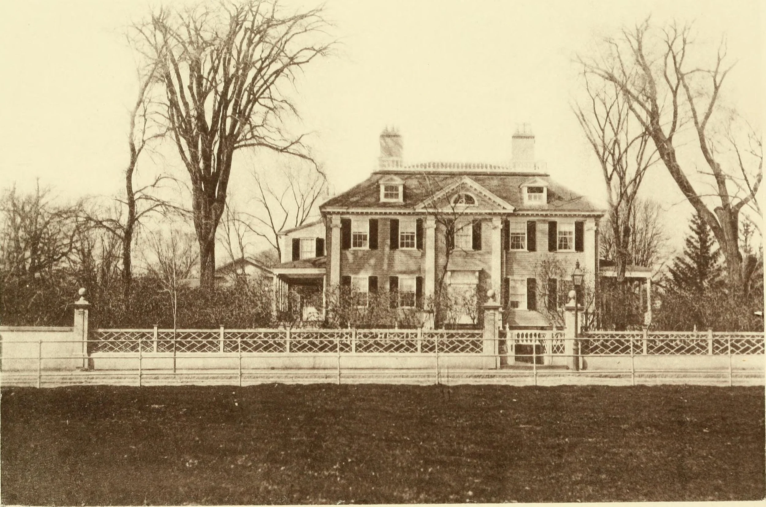 An old sepia photograph of a large, historic house with a portico, multiple chimneys, and several windows, surrounded by leafless trees and a decorative wooden fence in the foreground.