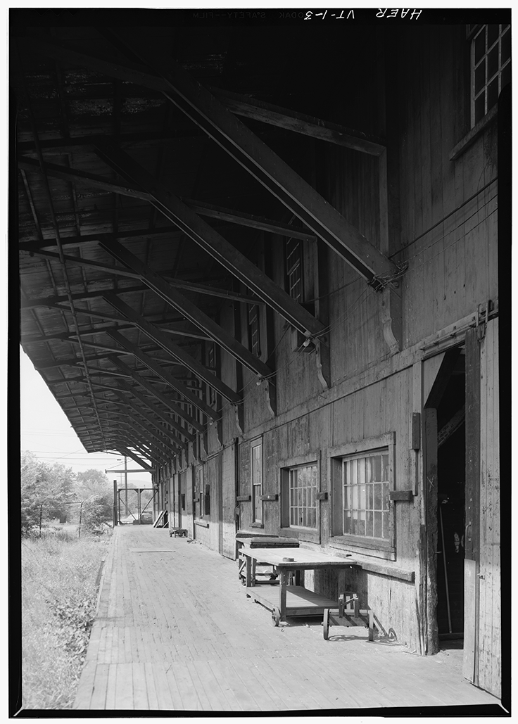 Black and white photo of an old wooden building with a covered porch, multiple windows, and some tables or workbenches outside.