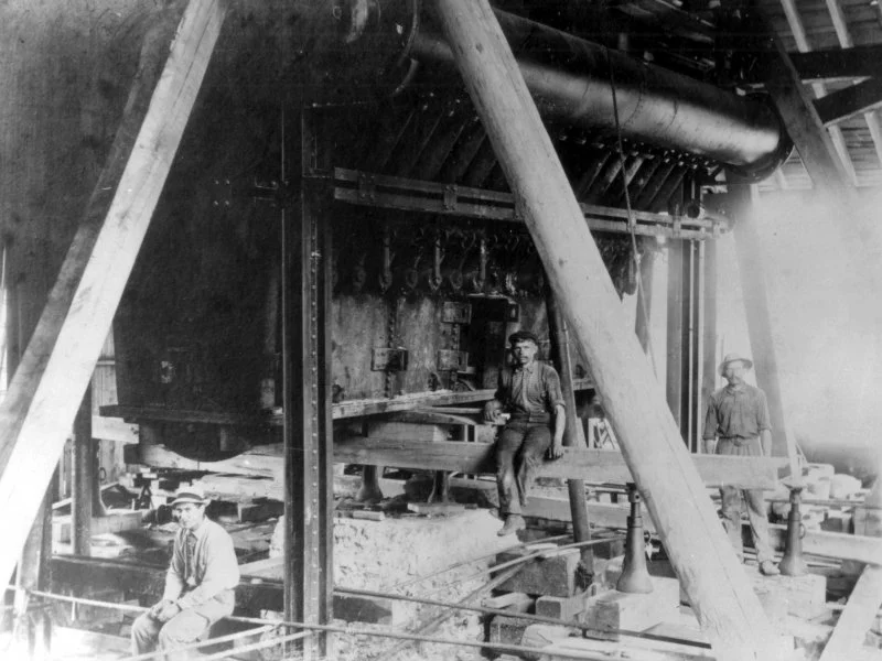 Black and white photo of three men working inside a large industrial workshop, possibly a sawmill or machinery factory, with large beams and machinery in the background.