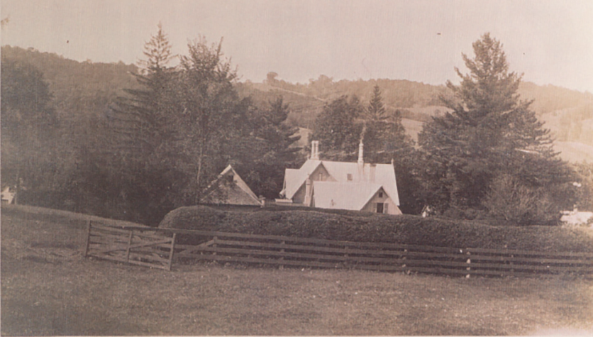 Old black-and-white photograph of a rural landscape with a house, a fence, trees, and hills in the background.