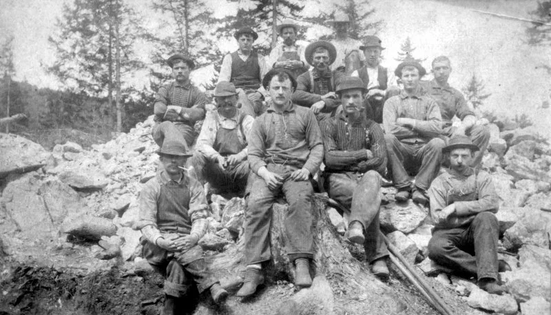 A black-and-white photo of eleven men sitting and standing outdoors on rocky terrain with trees in the background, some wearing hats and work clothes.