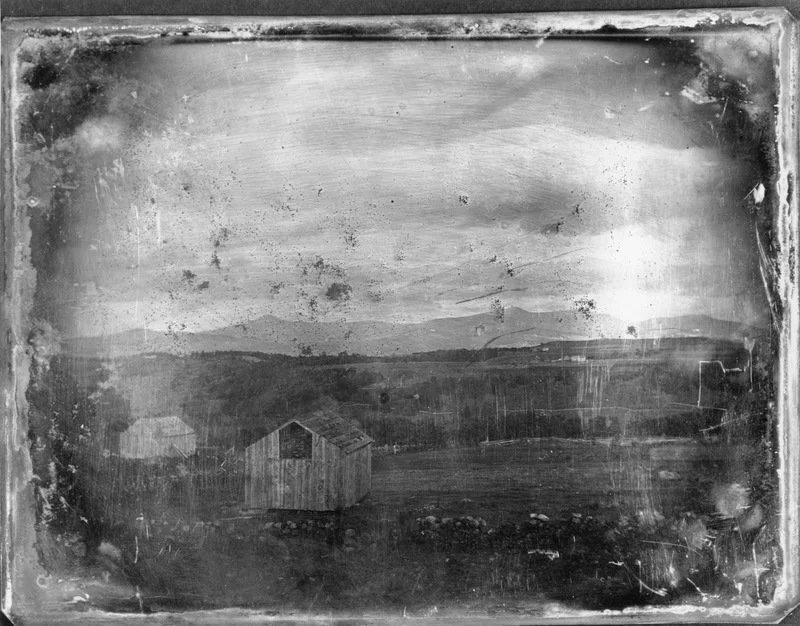 Black and white photo of a rural landscape with a small wooden barn, fields, hills in the distance, and a cloudy sky.