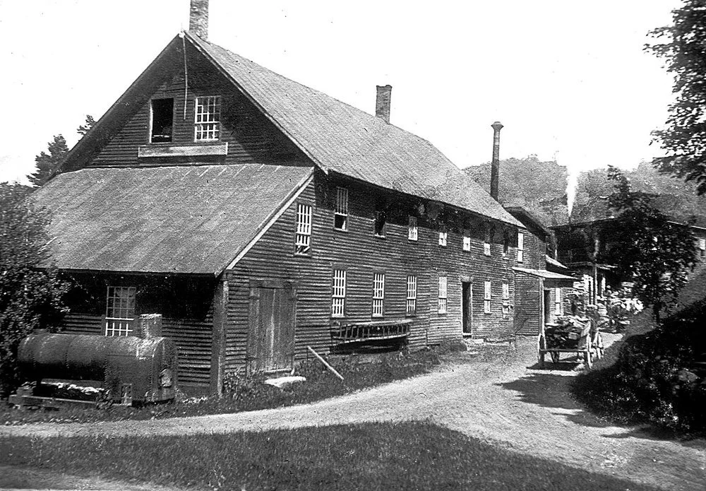 Old black-and-white photograph of a rustic wooden house with a sloped roof and multiple windows, surrounded by trees and a dirt path in the foreground.