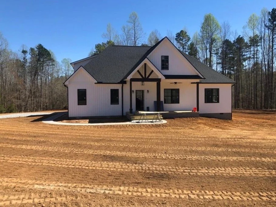 White house under construction with black trim on dirt lot, surrounded by trees, clear blue sky.
