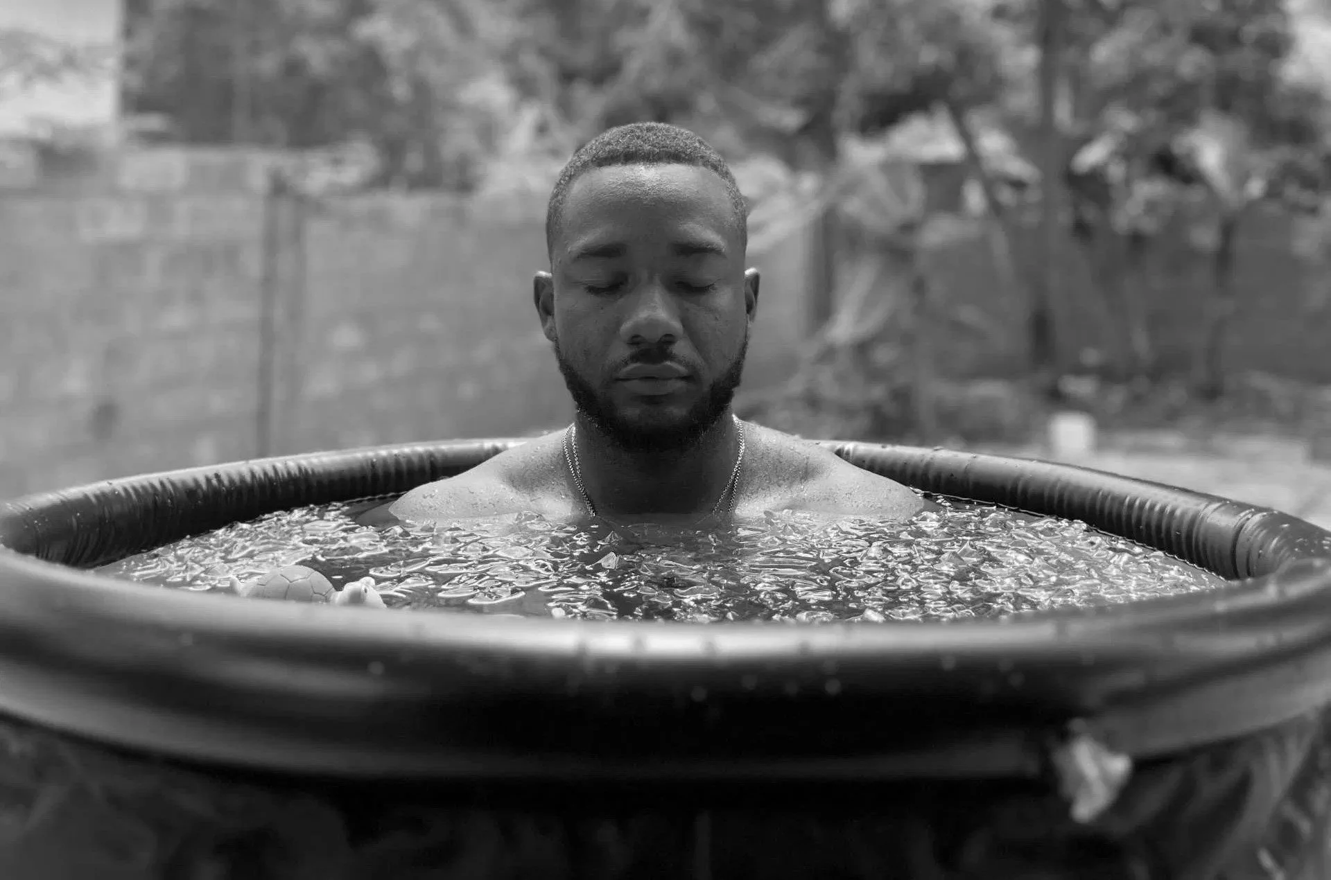 A young man with closed eyes and a beard relaxing in an outdoor hot tub, with trees and a fence in the background.