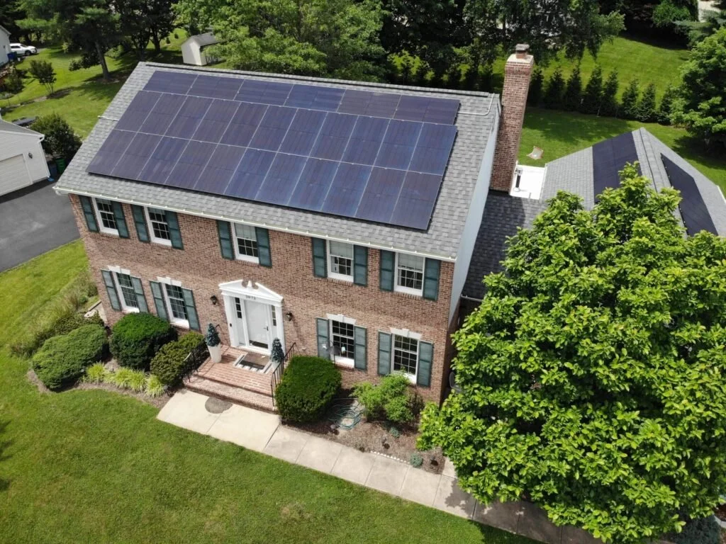 A two-story brick house with solar panels on the roof, a brick chimney, surrounded by green grass and trees, with a small front yard and a concrete walkway leading to the front door.