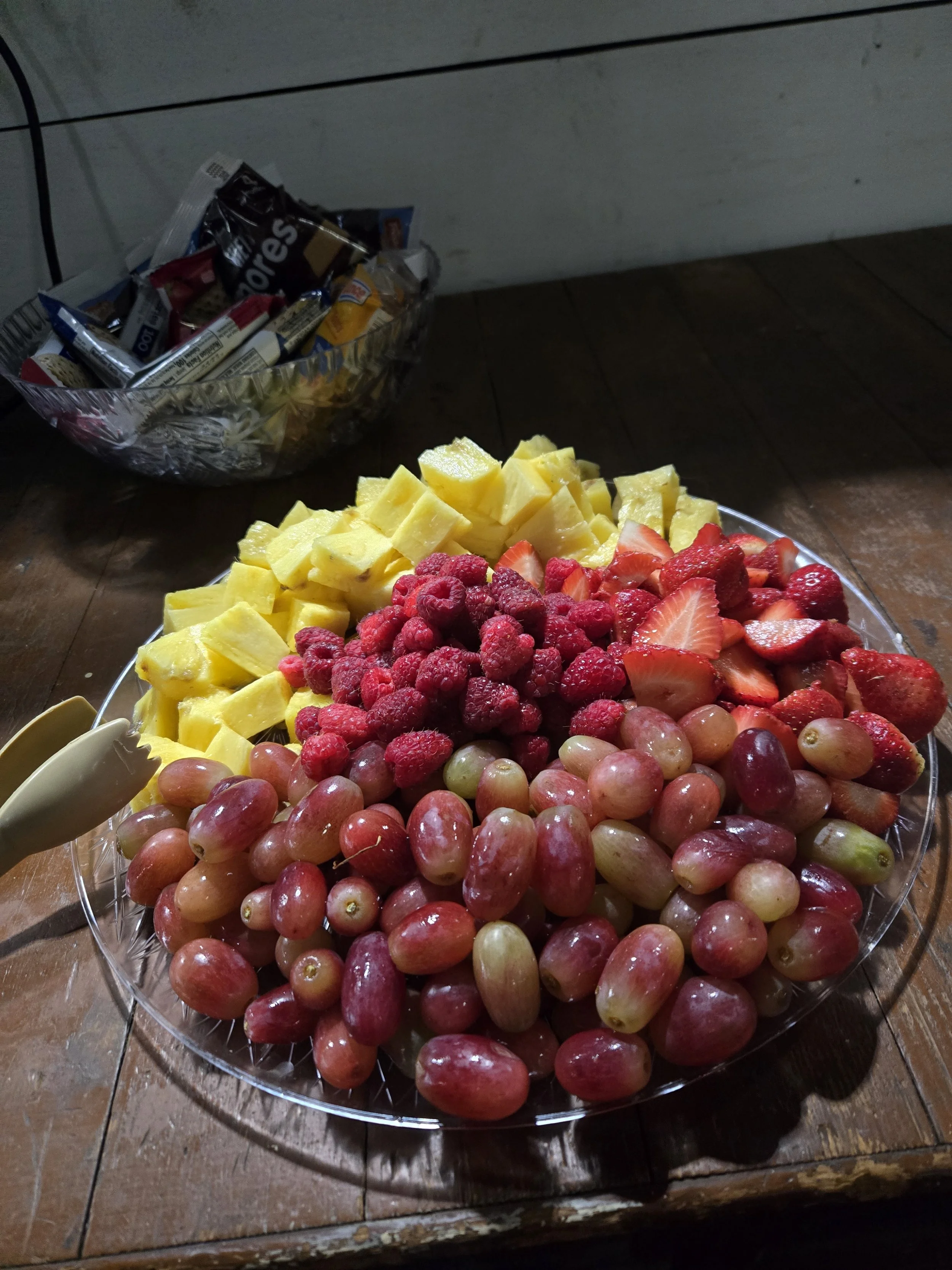 A large glass platter filled with grapes, strawberries, raspberries, and pineapple chunks. In the background, a bowl with snack packs on a wooden table.