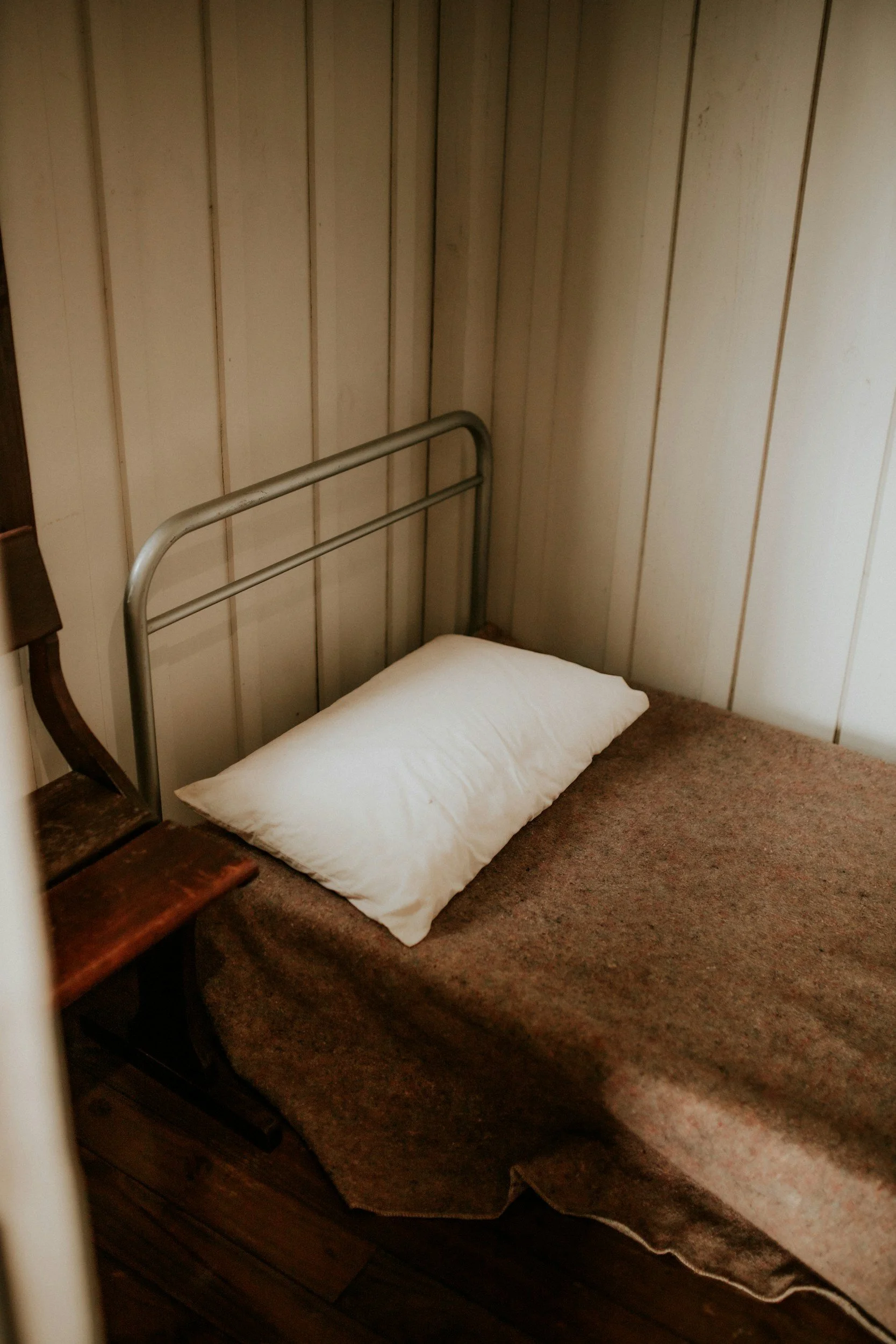 A simple bedroom with wooden paneled walls, a bed with a metal headboard, a brown blanket, and a single white pillow.